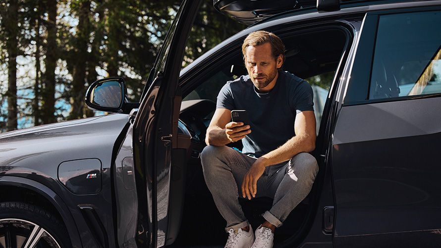 Mężczyzna siedzi w BMW patrzy na swój smartfon BMW Akcesoria Wiosenne i Letnie mężczyzna siedzący w BMW Patrzyący na swojego smartfona