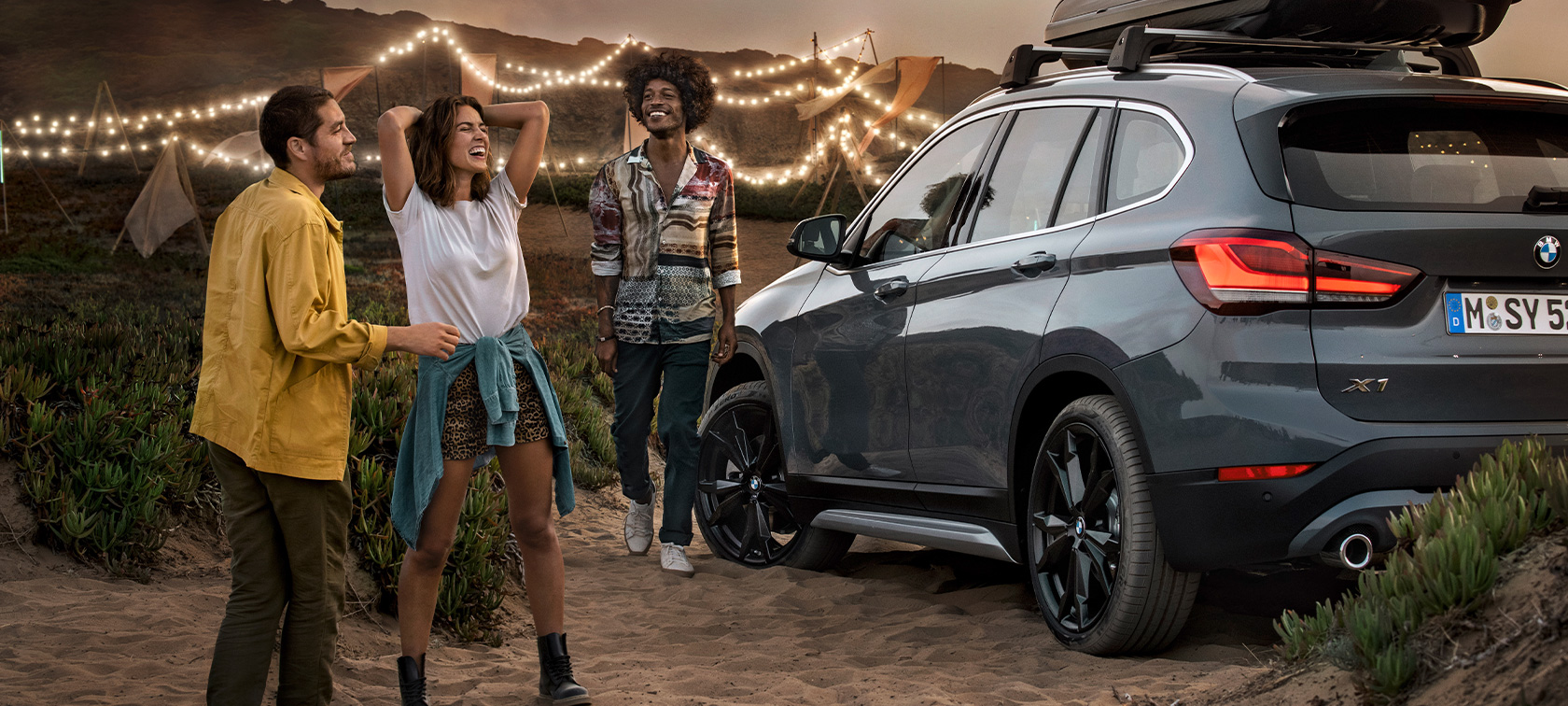 Szczęśliwe trzy osoby w BMW X1 z ofertą letnią BMW Szczęśliwe trzy osoby w BMW X1 w kolorze Mineral Grey z ofertą Letnią BMW widok trzy czwarte od tyłu na plaży Aksesoria BMW