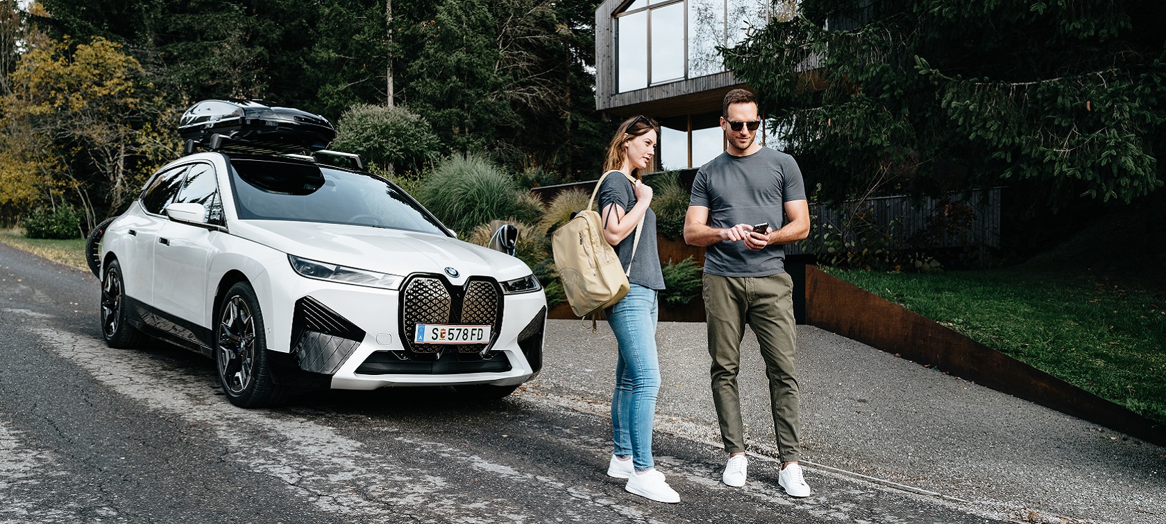 Mężczyzna z telefonem i kobieta z plecakiem stojący przed BMW iX Mężczyzna z telefonem i kobieta z plecakiem stojący przed BMW iX