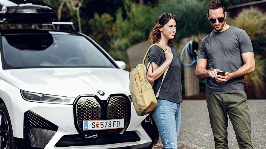 Mężczyzna z telefonem i kobieta z plecakiem stojący przed BMW iX Mężczyzna z telefonem i kobieta z plecakiem stojący przed BMW iX