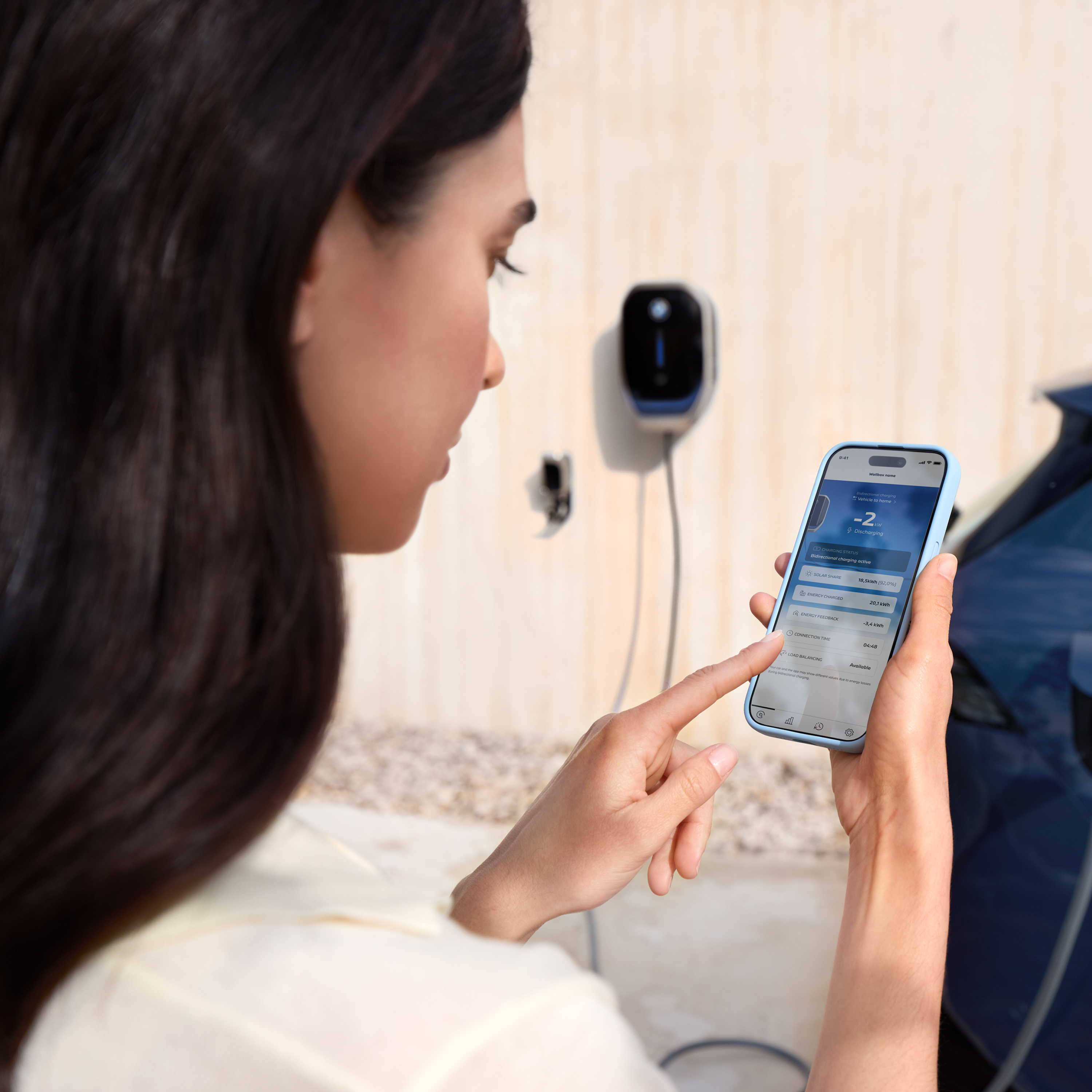 Kobieta patrzy na swój smartfon, a w tle jest stacja ładowania BMW Wallbox